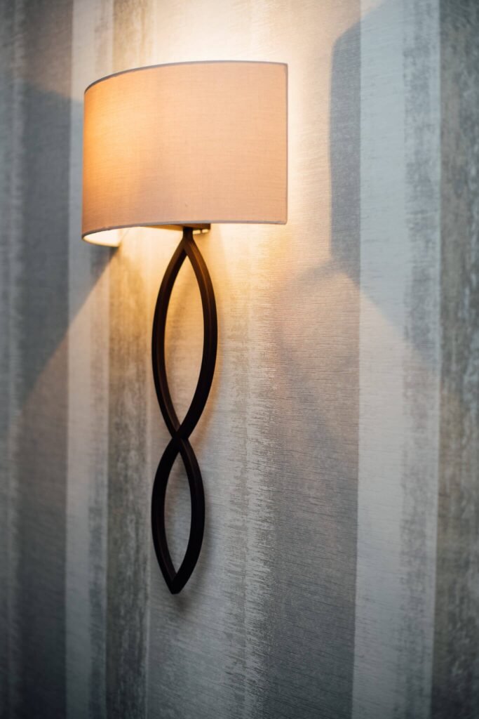 Modern wall sconce with a beige fabric shade, dark figure-eight base, illuminating striped textured wallpaper.