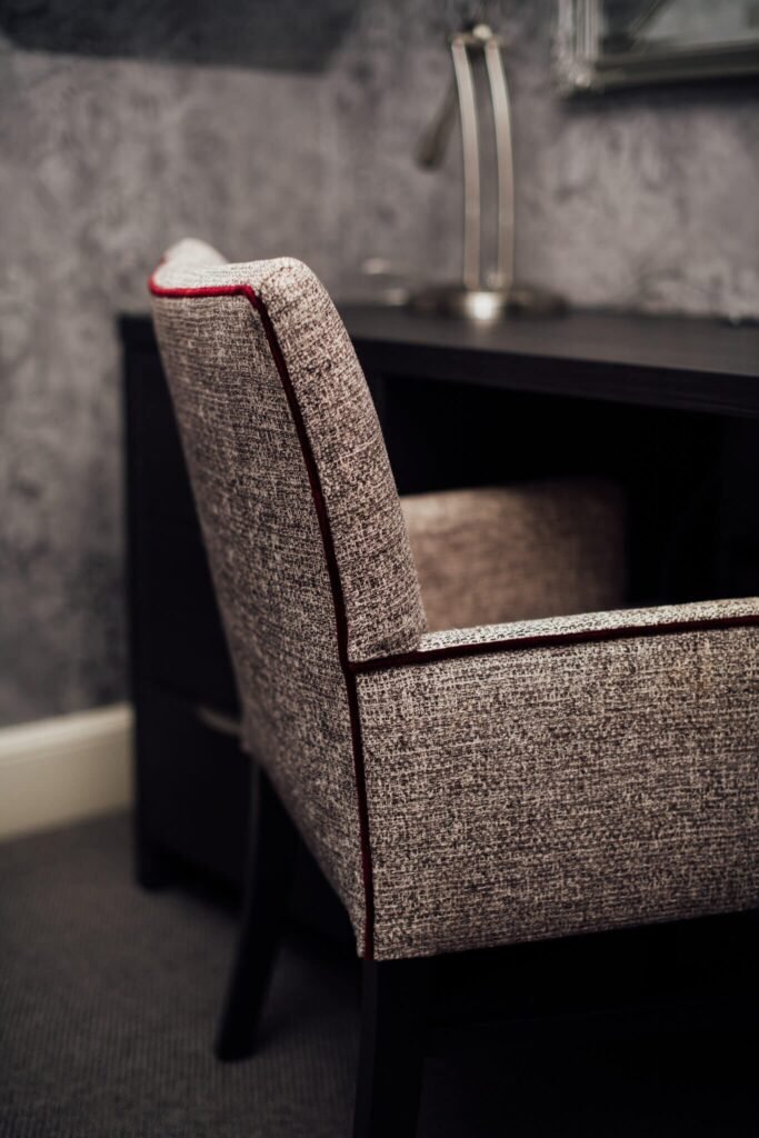 Textured gray chair with red piping, beside a dark desk with a silver lamp against a subtle textured wall.