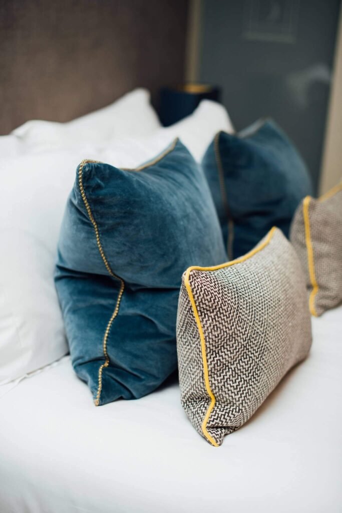 Decorative pillows on a bed: two blue velvet with gold beaded trim, two textured brown with yellow piping.