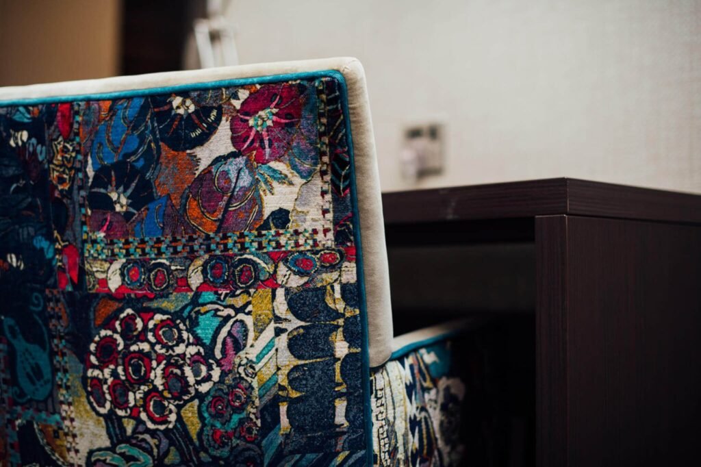 Close-up of a chair back with vibrant, intricate floral and abstract fabric, highlighted by turquoise piping, near dark wood furniture.