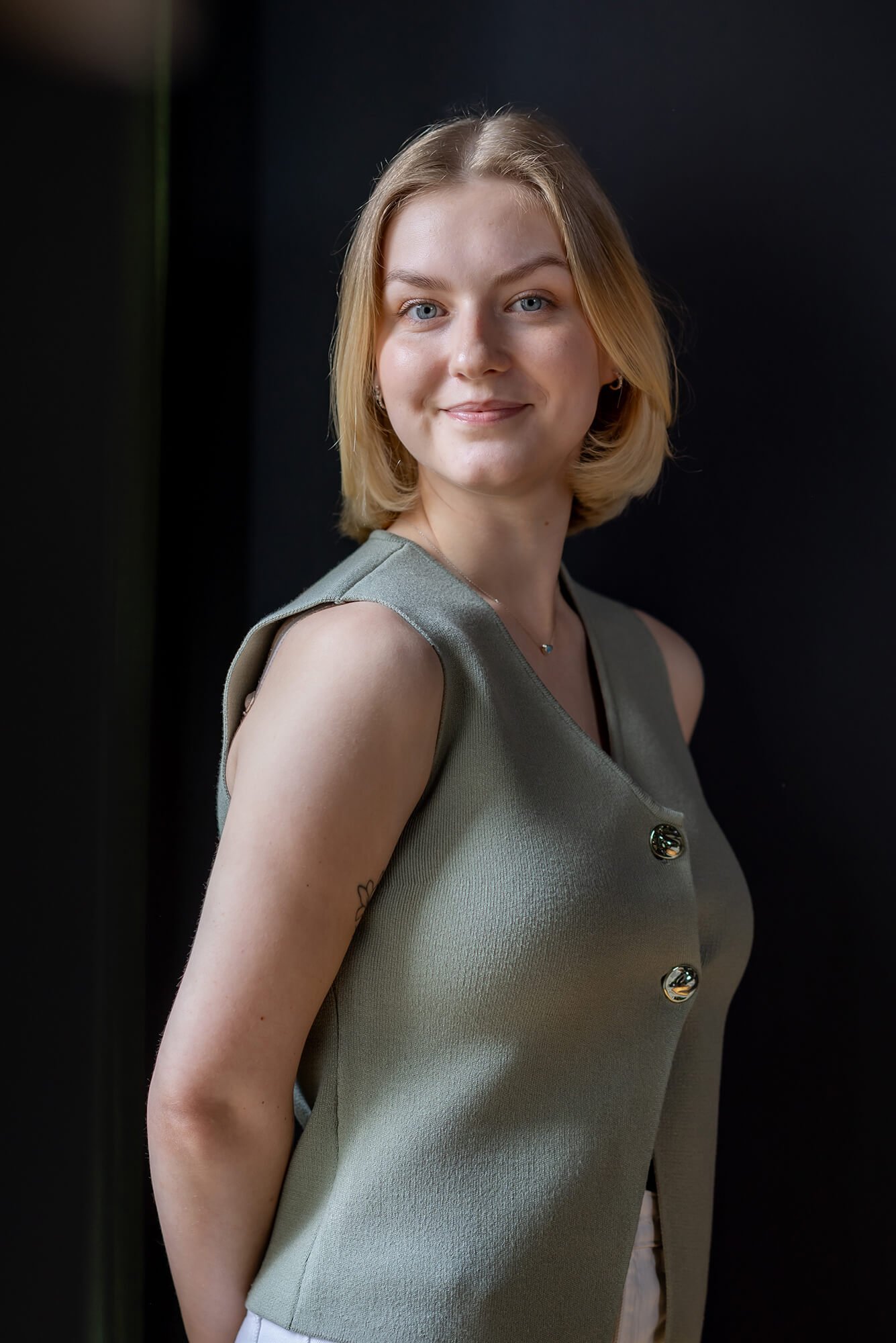 Smiling blonde woman with blue eyes, wearing a sage green sleeveless top with prominent gold buttons.