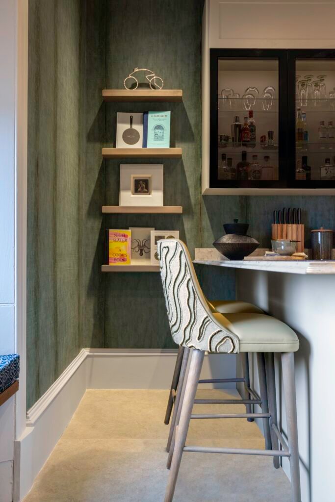 Elegant home bar featuring deep green textured walls, floating shelves with decor, marble counter, and stylish patterned bar stools.