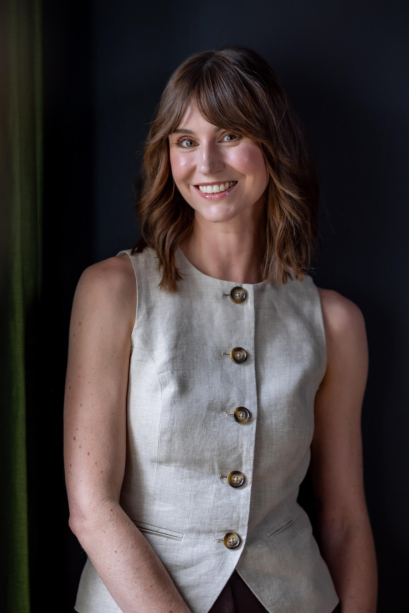 Smiling woman with shoulder-length brown hair in a light linen button-up top. Dark background with a vertical green curtain accent.