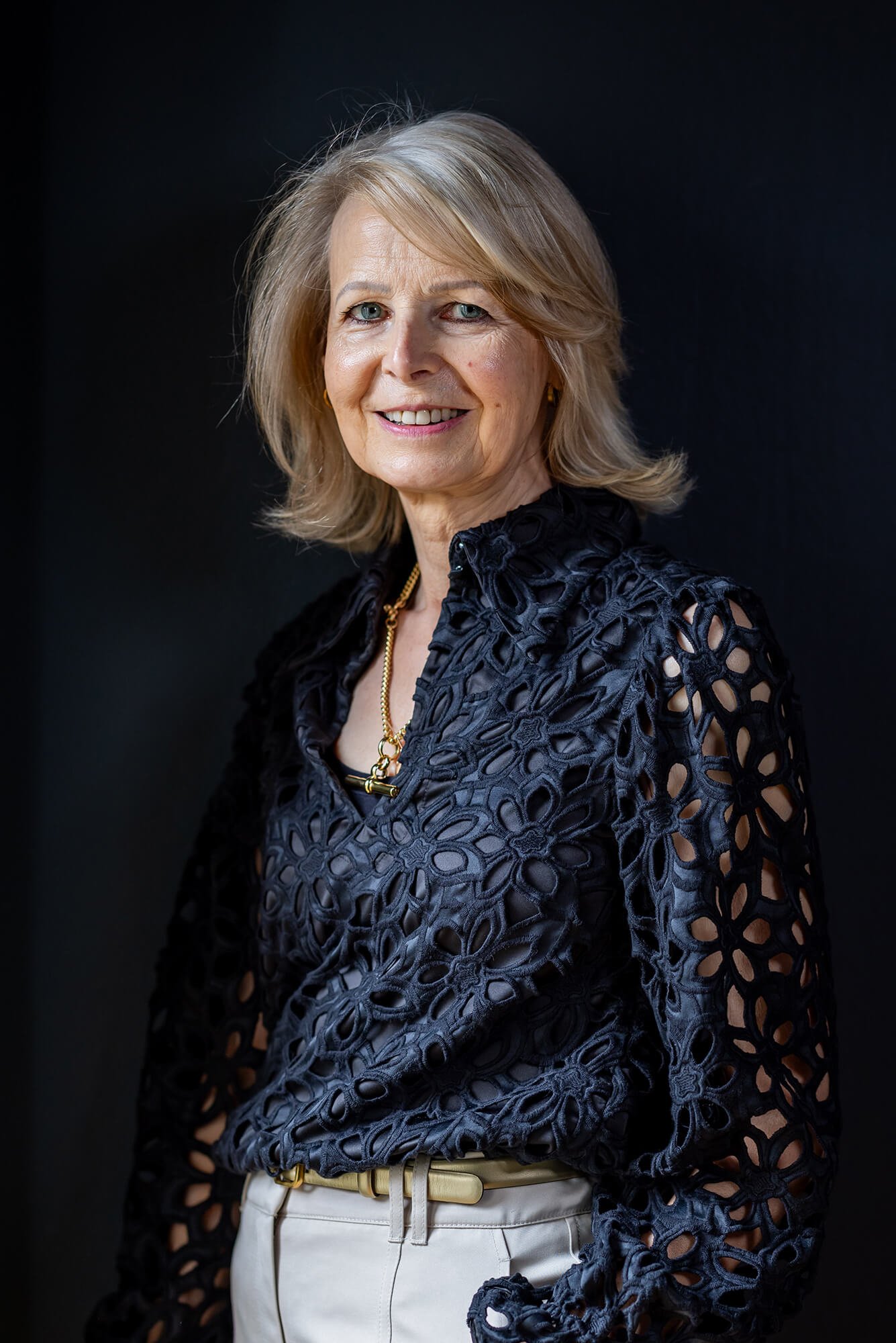 Smiling blonde woman in a black eyelet top, light pants, and gold necklace against a dark background.